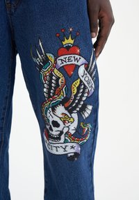 Jeans de mezclilla con un gran diseño bordado colorido que incluye una calavera, alas y un corazón, adornados con rhinestones y una cinta.