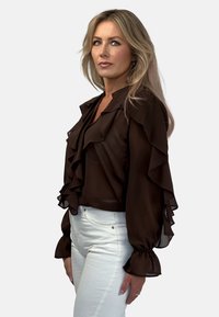 Brązowa przezroczysta bluzka z falbankami i długimi, balonowymi rękawami. Noszona z białymi, dopasowanymi dżinsami. Miękka tekstura, elegancki design.