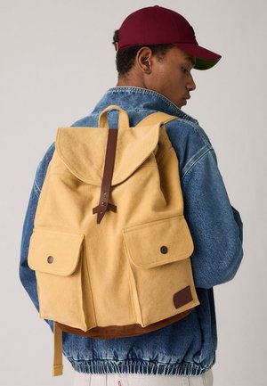 Joven con chaqueta de mezclilla y gorra roja mirando de perfil, llevando una mochila de lona beige con bolsillos frontales y detalles de correas de cuero.