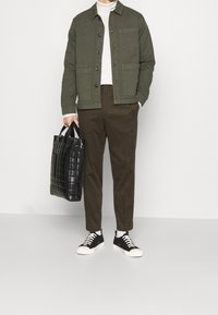 Olivgrön jacka med knappar, vit polotröja, bruna byxor, svart texturerad tote-väska och svarta sneakers med vita sulor.