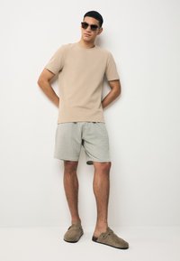 T-shirt beige à manches courtes, shorts rayés verts et blancs, chaussures plates en daim gris et lunettes de soleil foncées sur un fond neutre.