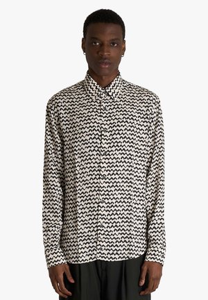 Uomo in piedi che indossa una camicia a maniche lunghe con motivo a zigzag bianco e nero e pantaloni neri su sfondo bianco semplice.