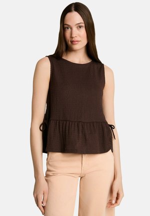 Mujer con cabello largo y castaño que lleva una blusa sin mangas de color marrón oscuro con textura y lazos laterales, y pantalones beige claro sobre un fondo liso.