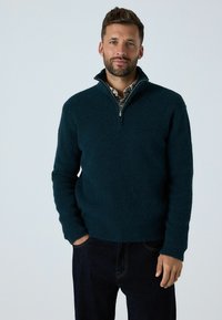 Pulover de polar verde închis cu guler cu fermoar pe un sfert, mâneci lungi și o suprafață texturată. Asortat cu pantaloni din denim închis.