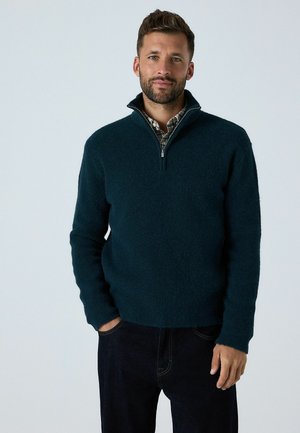 Dark teal fleece sweater with a quarter-zip collar, long sleeves, and a textured surface. Paired with dark denim pants.