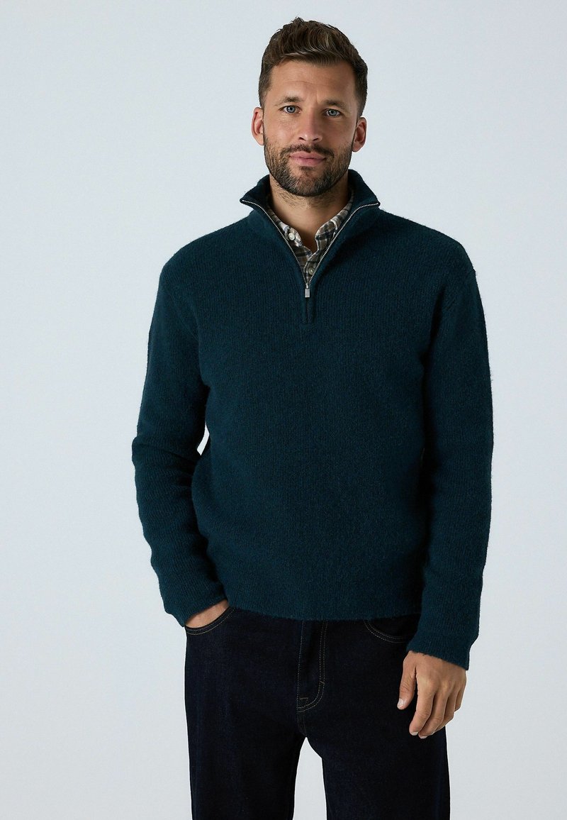 Pulover de polar verde închis cu guler cu fermoar pe un sfert, mâneci lungi și o suprafață texturată. Asortat cu pantaloni din denim închis.