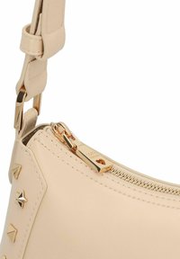 Beige leren handtas met een gouden rits, voorzien van piramidevormige studs langs de zijkant en een afneembare riem. Glad oppervlak, gebogen vorm.