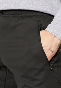 Schwarze Softshell-Hose mit einem Reißverschlusstaschen, glatter Textur und elastischem Bund. Eine Hand ist teilweise sichtbar, mit einem sichtbaren Tattoo.
