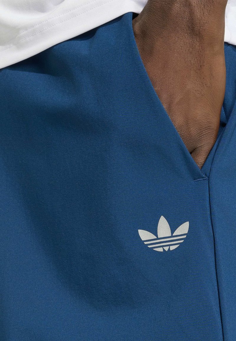 Dunkelhäutige Hand teilweise in der Tasche einer blauen Hose mit weißem Adidas-Trefoil-Logo neben der Tasche.