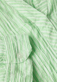 Tessuto a righe verde chiaro con una trama stropicciata, caratterizzato da un colletto abbottonato e un singolo bottone bianco sul lato sinistro.