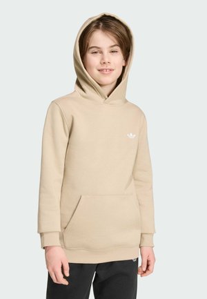 Ung person smilende, iført en beige hættetrøje med kængurulomme og sorte bukser, stående mod en ensfarvet lys baggrund.