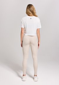 Witte cropped T-shirt met korte mouwen en een klein logo op de achterkant, gecombineerd met beige leggings en witte sportieve schoenen.