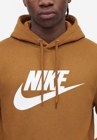 Κοντινό πλάνο ατόμου που φορά καφέ φούτερ Nike με κορδόνια και μεγάλο λευκό λογότυπο Nike στο στήθος.