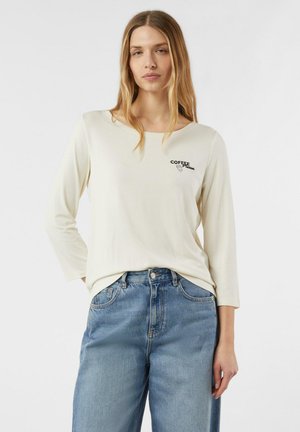 Cremefarbener Langarmshirt mit rundem Ausschnitt und schwarzem "COFFEE"-Schriftzug. Kombiniert mit hellblauen, weit geschnittenen Denim-Jeans.