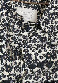 Chemise avec motif floral noir sur tissu blanc, boutons noirs et une étiquette indiquant « PART TWO, Established 1986, Copenhagen, 36. »