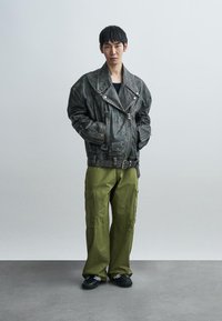 Μαύρο δερμάτινο μπουφάν μοτοσικλετιστή με μεγάλο γιακά, φερμουάρ και ζώνη, συνδυασμένο με πράσινο παντελόνι cargo και μαύρα παπούτσια.