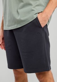 Black cotton shorts with pockets, featuring a stitched hem and a small logo detail. Worn with a light green t-shirt.