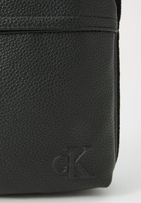 Black textured leather shoulder bag featuring a debossed logo, flat design, and subtle seam details. Compact and functional.