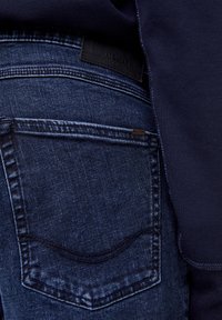 Mörkblå denim jeans med en något urtvättad yta. Har en bakficka och en svart märkeslapp. Intill en marinblå tyg.