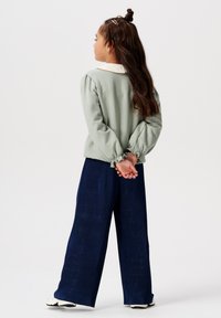 Jong meisje met lang bruin haar, gekleed in een lichtgroene blouse met witte kraag en marineblauwe geplooide wijde broek, staande met de handen achter de rug.