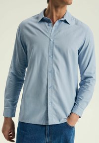 Jasnoniebieska koszula z długim rękawem i kołnierzykiem zapinanym na guziki, z niebieskimi guzikami i miękką fakturą, noszona z ciemnymi dżinsami.