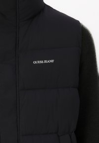 Black padded vest with high collar, featuring a white "GUESS JEANS" logo on the chest. Smooth texture with quilted detailing.