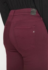 Vaqueros skinny burdeos hechos de tejido elástico, con un bolsillo trasero que presenta una discreta costura en Chevron y una etiqueta de cuero negro en la cintura.