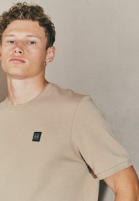 Camiseta de algodón beige de manga corta, con un pequeño parche de logo negro en el pecho. Textura suave con un diseño clásico de cuello redondo.