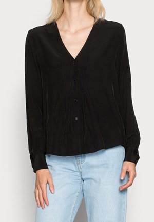 Woman wearing a black buttoned V-neck blouse with long sleeves and light blue jeans, standing against a plain background.