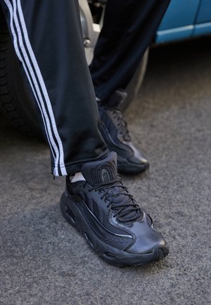 Personne portant des baskets noires à semelles épaisses et un pantalon de survêtement noir avec des bandes blanches latérales, debout près d'un pneu de véhicule bleu.