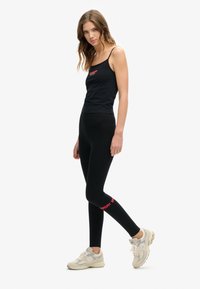 Top nero senza maniche con spalline sottili, abbinato a leggins neri con testo rosso "Superdry". Il modello indossa scarpe da ginnastica di colore chiaro.