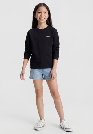 Dziewczyna uśmiechająca się, ubrana w czarną bluzę Calvin Klein, jasnoniebieskie dżinsowe szorty i białe trampki, stoi na jednolitym tle.