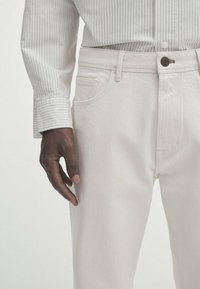 Valkoiset denimin farkut ovat suoraa leikkausta, sivutaskuilla ja edessä nappikiinnitys. Kangas on sileäpintainen, ja sitä täydentää vaaleanvärinen, ohutraidallinen paita.