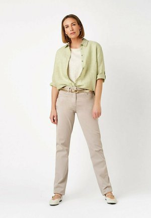 Donna in piedi che indossa una camicia verde chiaro con bottoni sopra una canotta bianca, pantaloni beige con cintura e scarpe basse bianche, su sfondo bianco.