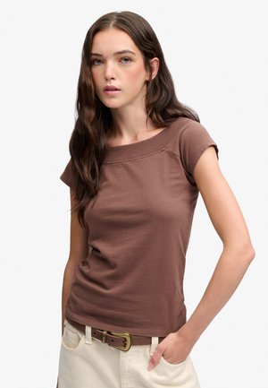 Giovane donna con lunghi capelli scuri indossa una maglia marrone a maniche corte e pantaloni beige con una cintura marrone, posando con una mano in tasca.