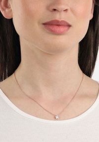 Donna che indossa una delicata collana in oro rosa con un piccolo pendente a forma di fiore incastonato con diamanti, abbinata a un top bianco a costine.