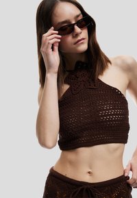 Top corto de punto marrón con un diseño floral en el escote y textura de tejido abierto. La modelo lleva gafas de sol oscuras, mostrando un estilo minimalista.