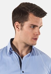 Camicia a scacchi blu e bianca con colletto aderente, polsini interni navy a contrasto e bottoni neri. Texture in cotone morbido.
