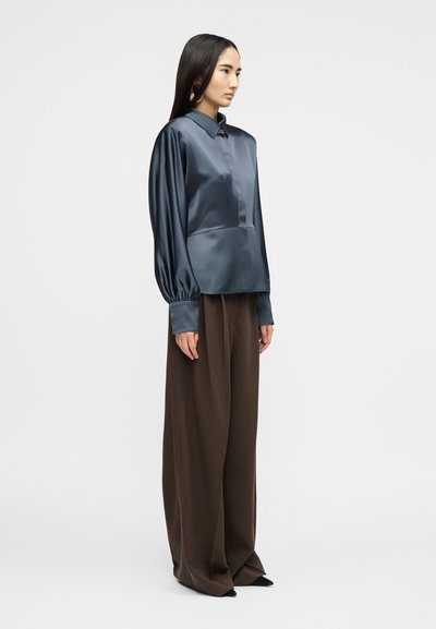 Γυναίκα με μακριά σκούρα μαλλιά στέκεται πλάγια φορώντας μια λαμπερή μπλε σκούρα σατινέ μπλούζα με φουσκωτά μανίκια και φαρδιές καφέ παντελόνες.