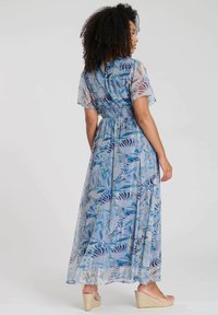 Robe maxi en tissu bleu clair avec impression botanique. Manches courtes, taille froncée, jupe fluide, portée avec des sandales compensées beiges.
