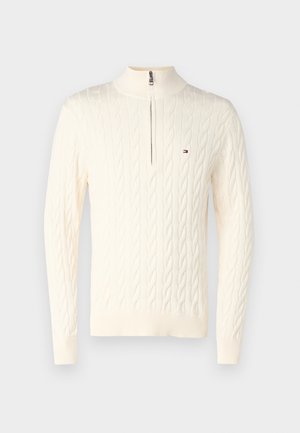 Suéter crema de punto trenzado con un alto cuello y una cremallera corta. Presenta puños y dobladillo acanalados, con un pequeño logo en el pecho.