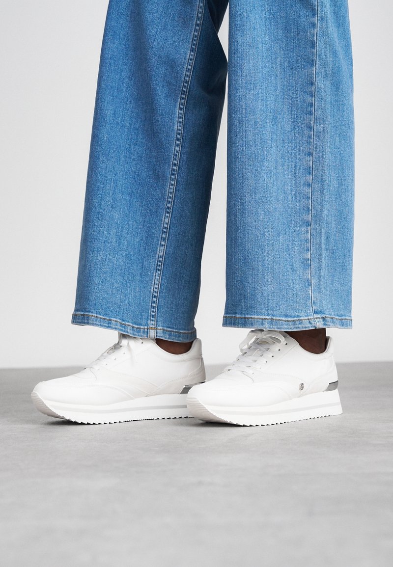 Personne portant un jean bleu à jambes larges et des baskets blanches à plateforme, debout sur une surface grise.