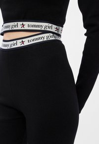 Crne tajice sivi pojas s natpisom "tommy girl" i crvenom zvijezdom. Glatka tekstura, prilagođen dizajn. Djelomičan prikaz.