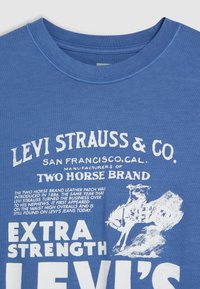 Tricou din bumbac albastru, cu text și grafica imprimate în alb, incluzând un cal și un călăreț, cu guler rotund și mâneci scurte.
