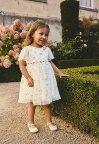 Weißes Kleid mit floraler Stickerei und kurzen Ärmeln, mit mehrlagigem Tüllrock. Das Outfit wird durch hellrosa Ballerinas ergänzt.