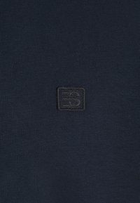 T-shirt bleu marine en tissu côtelé doux arborant un petit logo embossé dans le coin supérieur gauche. Design simple sans motifs additionnels.