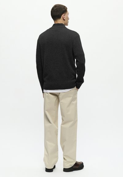 Pull en tricot gris foncé avec un col montant, associé à un pantalon large couleur crème. Les chaussures présentent des accents marron foncé avec une semelle texturée.
