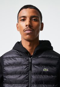 Sort puffer-vest med horisontale sømmer, frontlynlås, og Lacoste-logo. Båret over en svart hettegenser med en glatt, blank tekstur.