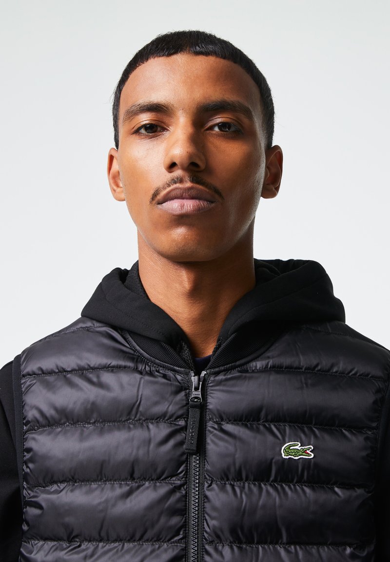 Gilet rembourré noir avec des coutures horizontales, fermeture éclair sur le devant et un logo Lacoste. Porté par-dessus un sweat à capuche noir, avec une texture lisse et brillante.