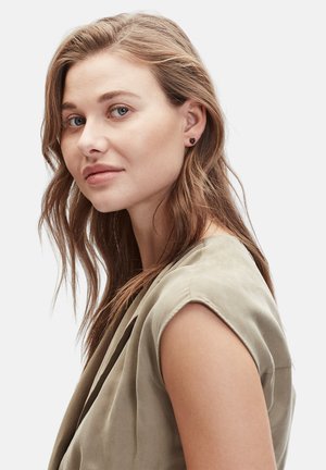 Haut sans manches de couleur neutre en tissu doux et fluide ; le modèle a de longs cheveux ondulés et de petites boucles d'oreilles en forme de cœur noir.
